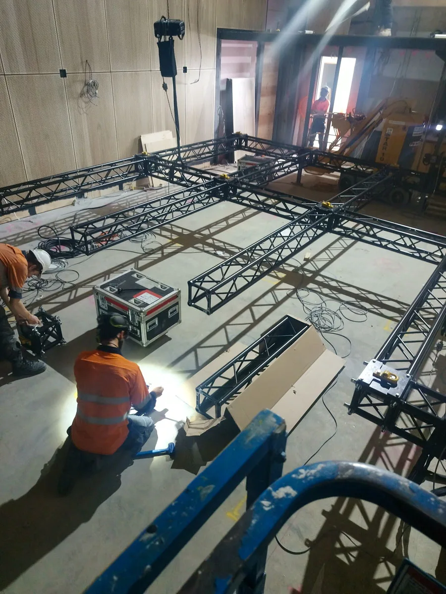 Crew assembling a box-truss pipe grid with a chain hoist overhead at ECU